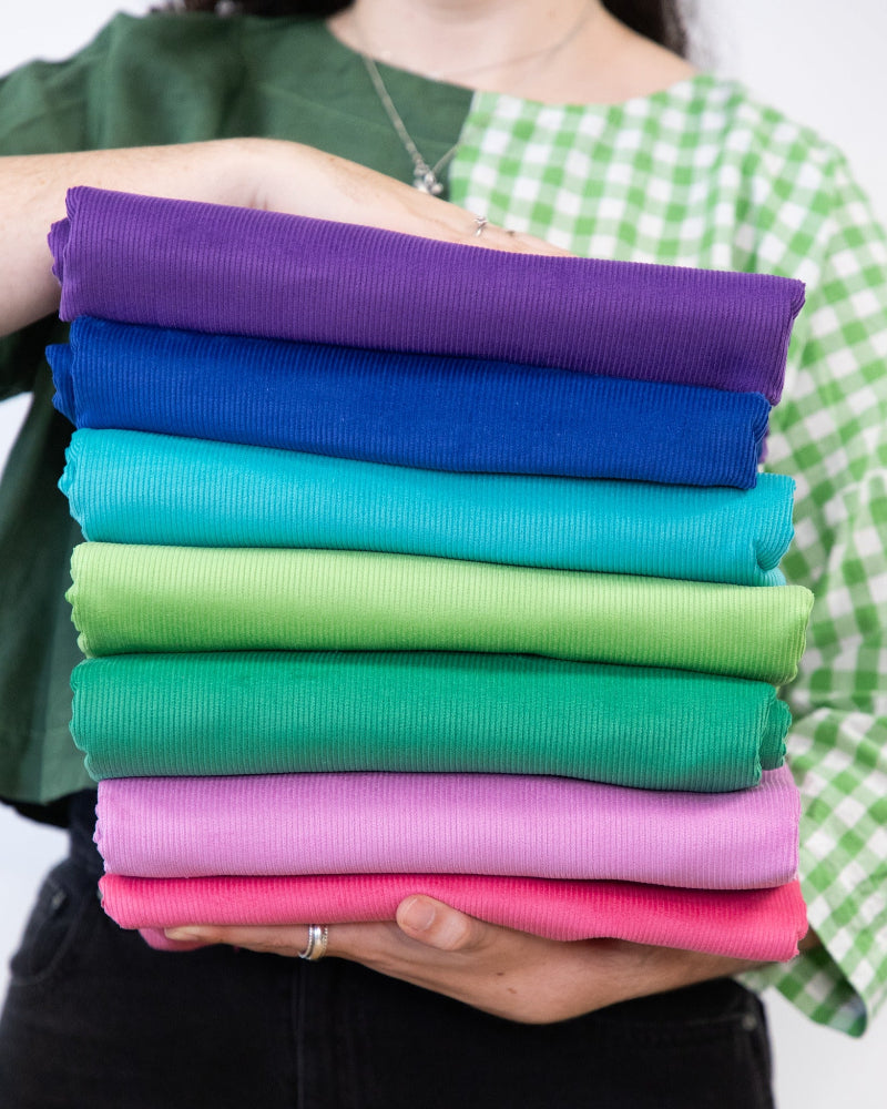 A stack of eight folded corduroy fabrics in purple, blue, teal, lime green, forest green, light pink, and hot pink, held by a person wearing a green and white checkered shirt.
