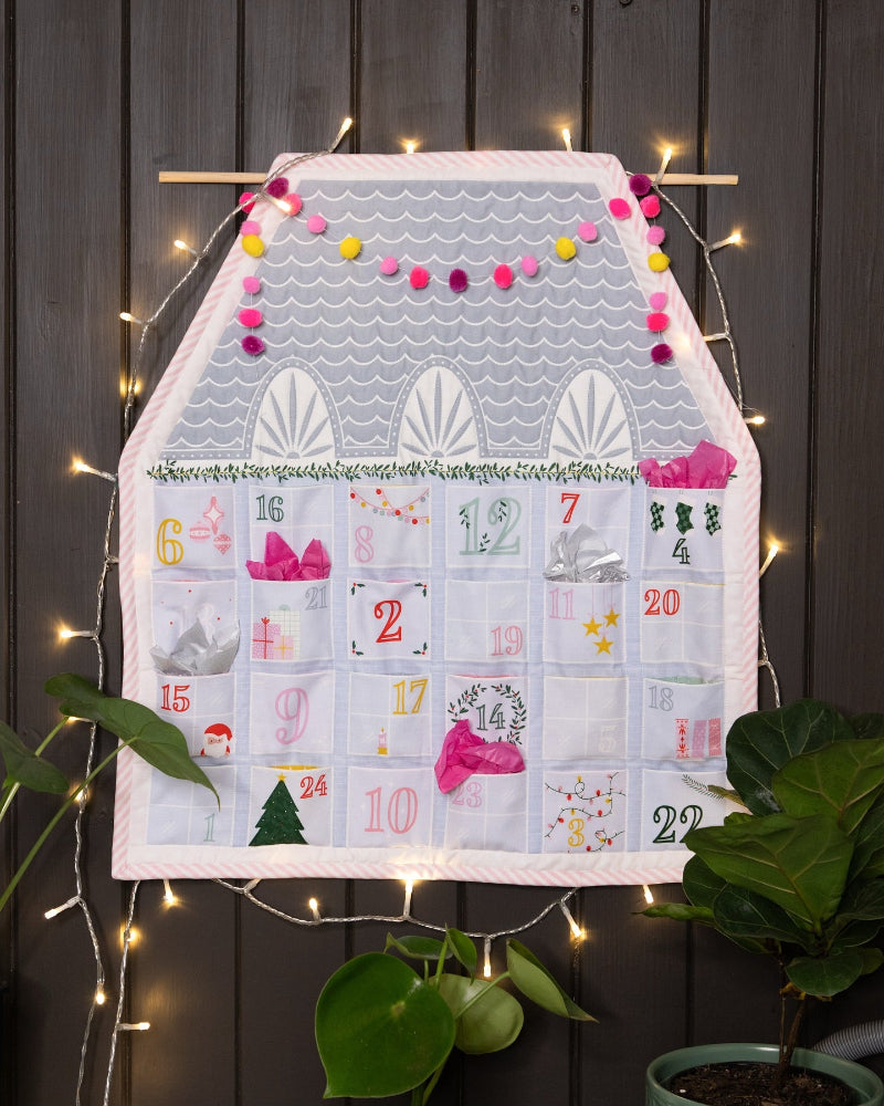 A house-shaped Advent calendar hanging on a dark wood paneled wall, decorated with warm fairy lights and green potted plants. The calendar features a white and gray wavy pattern top, a pink garland, and multiple numbered white pockets with festive illustrations and text.