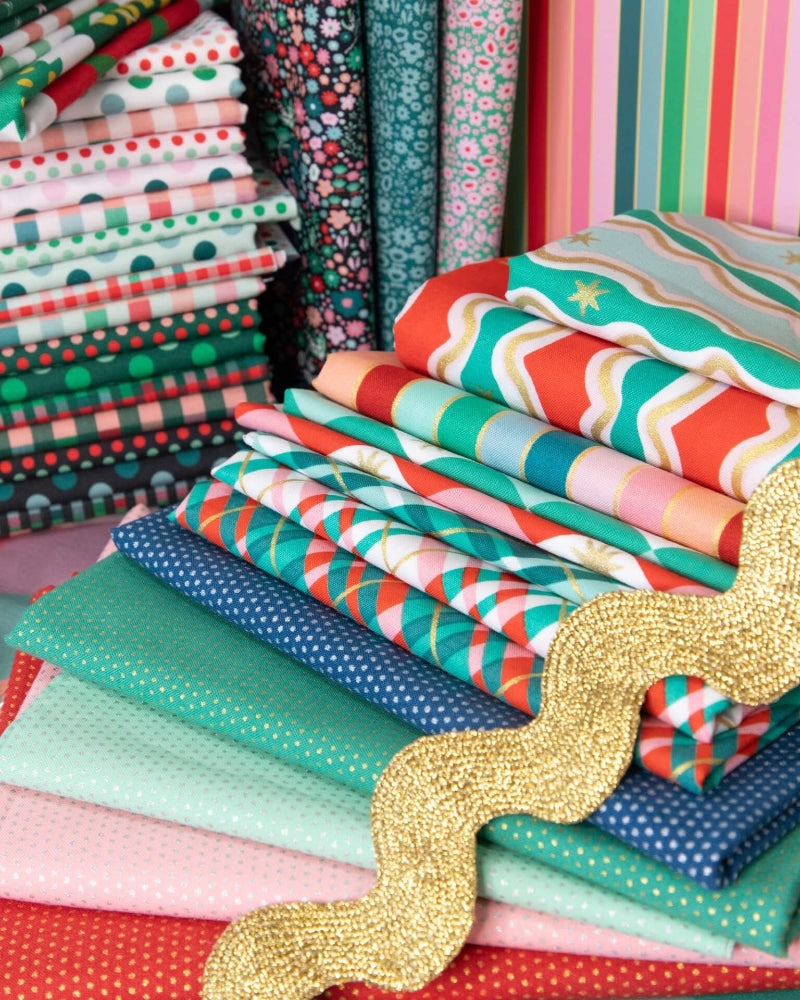 A stack of colorful patterned fabric bolts in various designs including stripes, polka dots, florals, and plaid, arranged with a glittery gold wavy ribbon in the foreground.